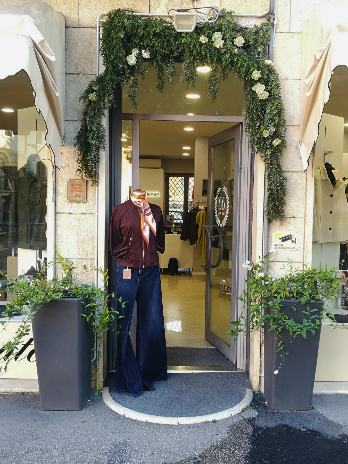 Ingresso esterno del negozio Danese Rinaldo: porta aperta con manichino in giubbotto bordeaux, ghirlanda di rose bianche e targa Bottega Storica di Verona